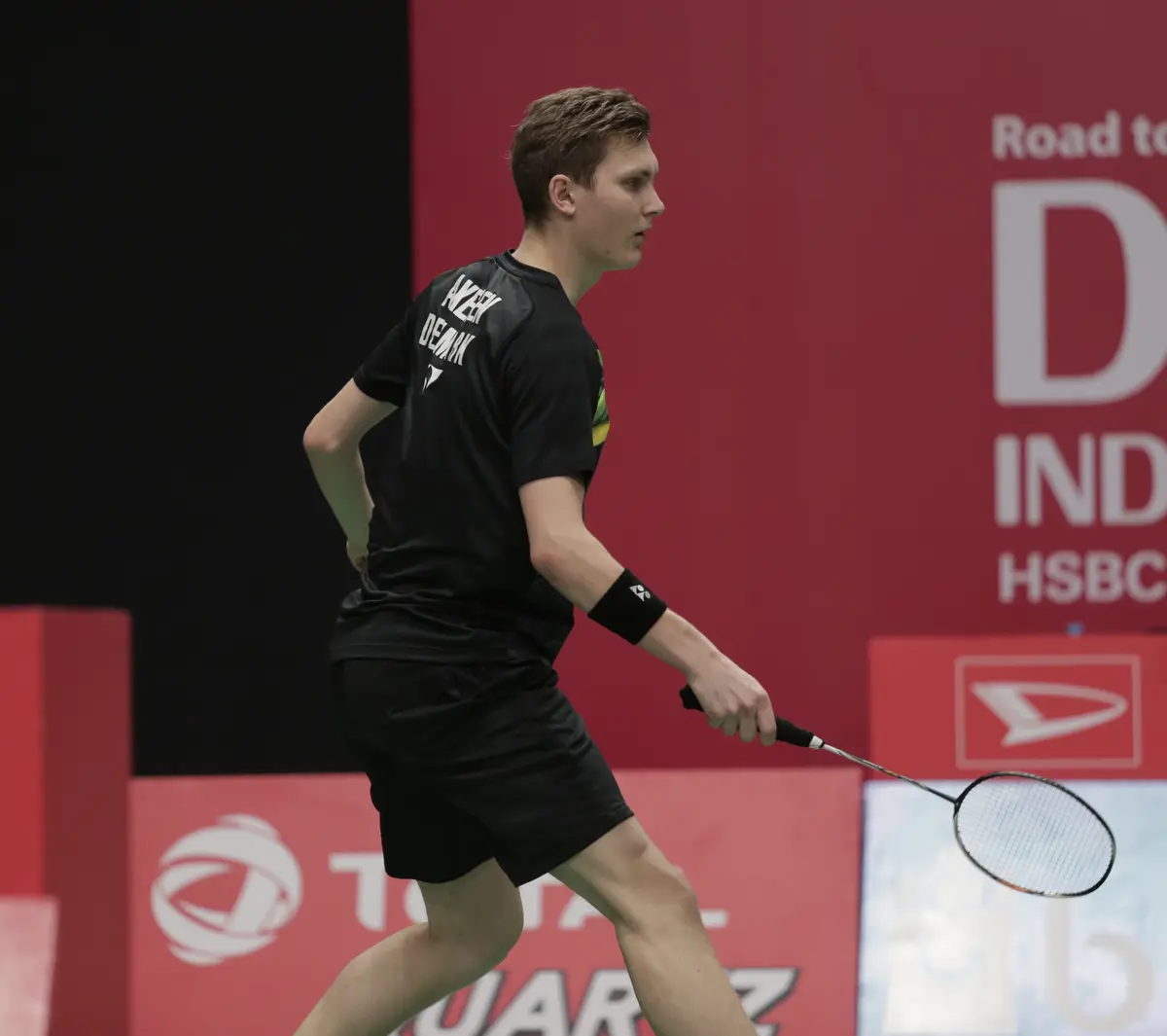 Viktor Axelsen, en maillot et short noirs avec un bracelet au poignet, tient sa raquette de badminton le long du corps sur un court, devant des panneaux publicitaires rouges.