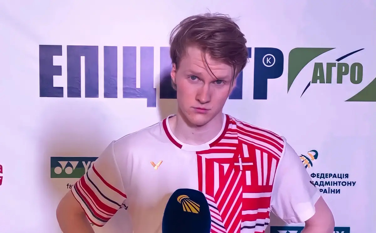Un joueur de badminton aux cheveux châtains, en maillot blanc et rouge à motifs, répond à une interview devant un panneau publicitaire, un micro jaune et bleu au premier plan.