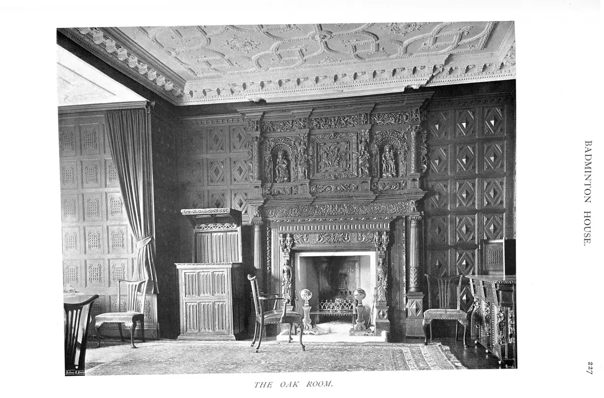 Photographie ancienne en noir et blanc d'un salon lambrissé de chêne, avec une grande cheminée sculptée, un plafond à décor de stucs, des chaises et un meuble en bois.