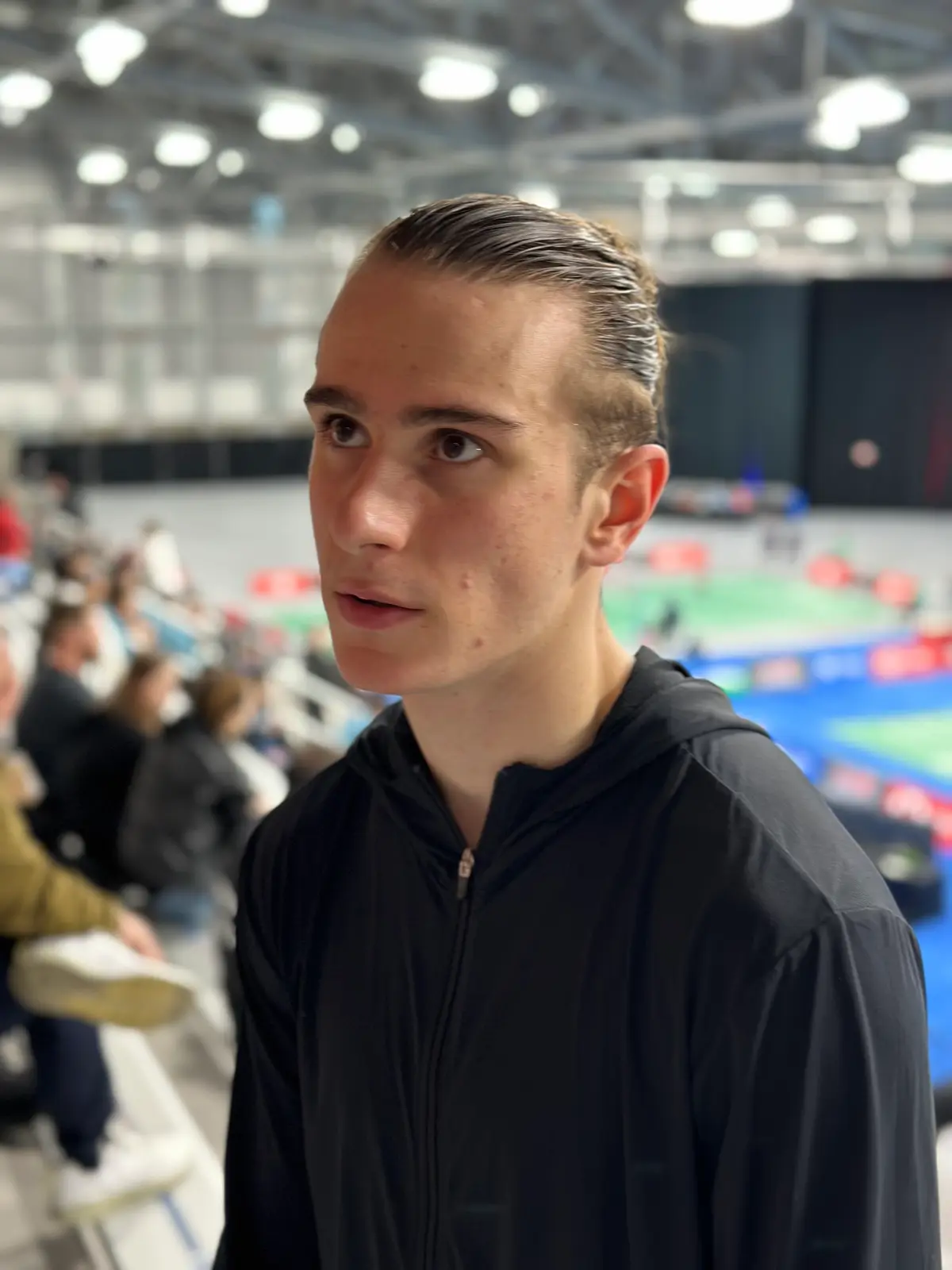 Alex Lanier en haut noir à capuche, de face dans les gradins d’une salle où l’on distingue des terrains de badminton flous.