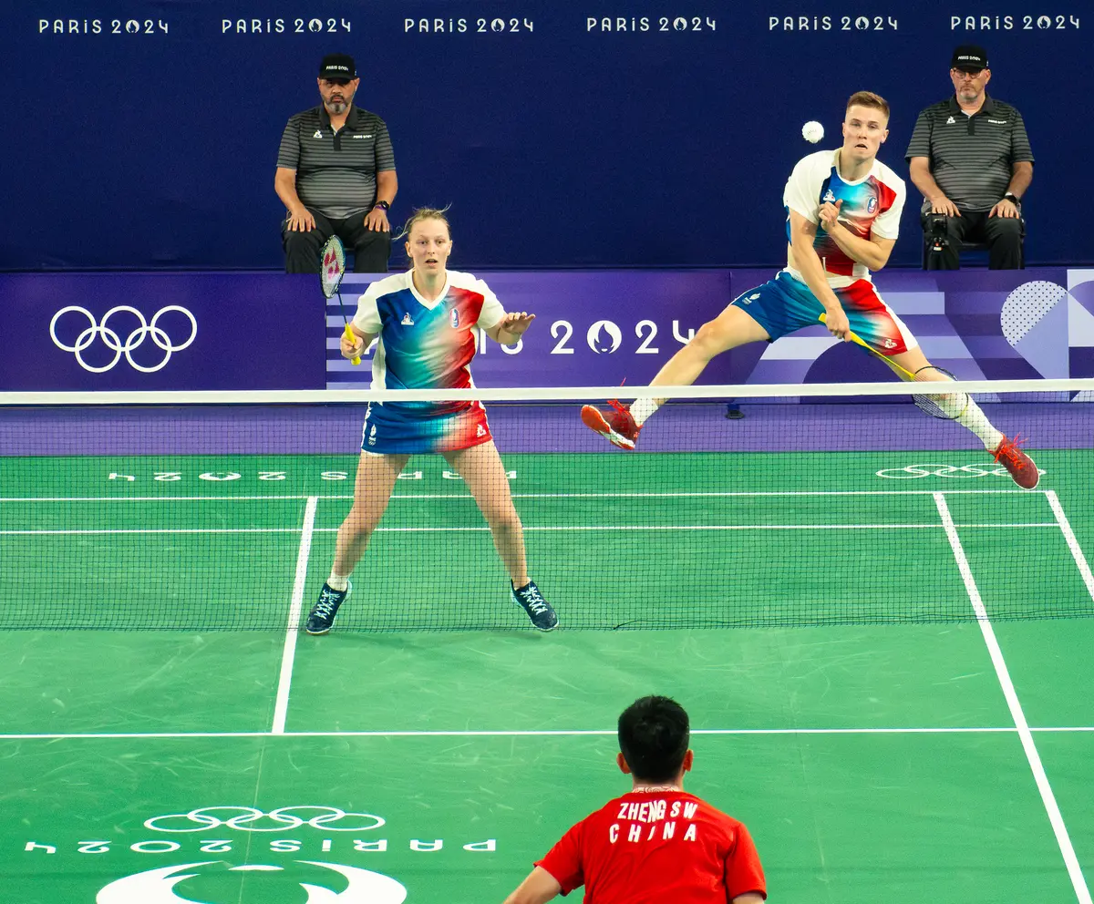Un joueur en tenue bleu, blanc et rouge saute près du filet pour frapper le volant, pendant que sa partenaire attend en position, face à un adversaire en rouge, lors d'un match de badminton aux Jeux de Paris 2024.