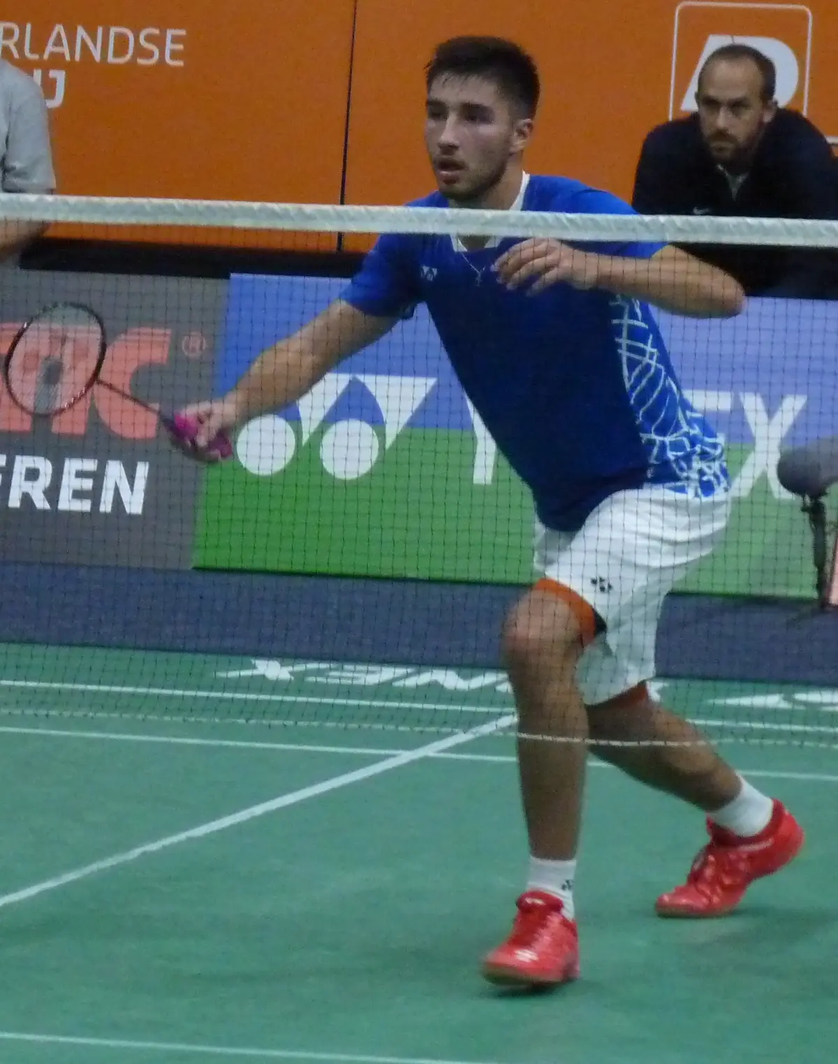 Toma Junior Popov, en maillot bleu et short blanc, fléchit les jambes près du filet et tend sa raquette sur un terrain de badminton vert, devant des panneaux publicitaires orange.