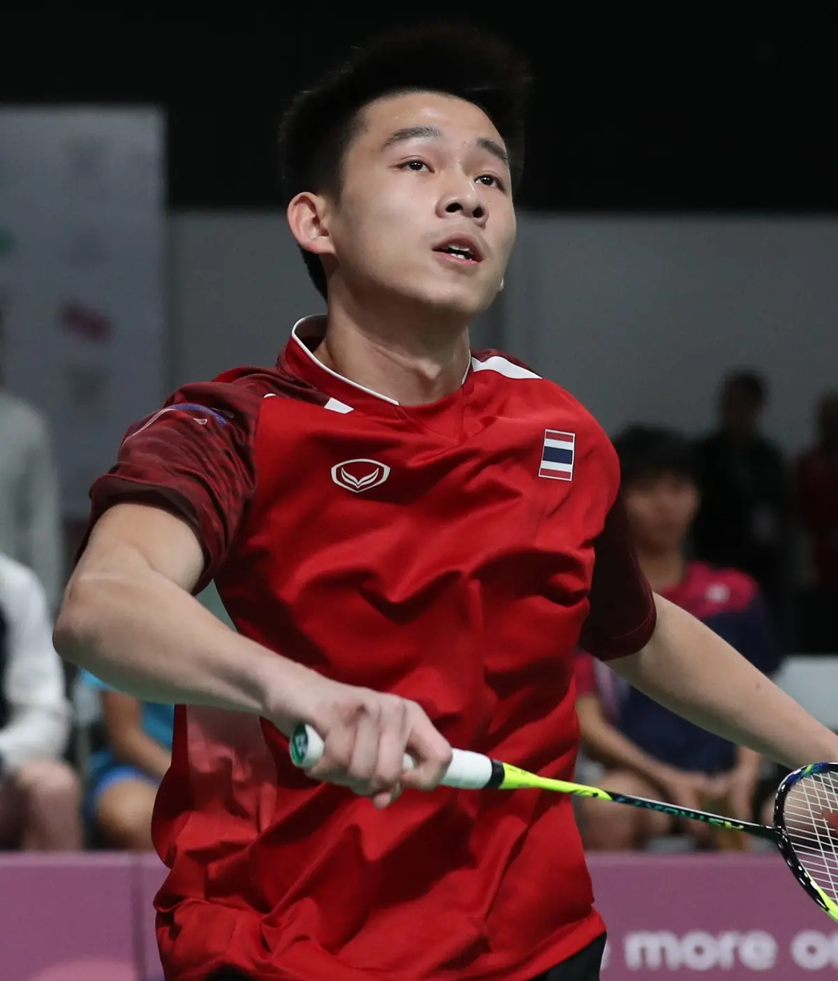 Un joueur de badminton en maillot rouge orné d'un drapeau thaïlandais, le regard levé, tient sa raquette à hauteur de la taille dans une salle avec du public flou en arrière-plan.