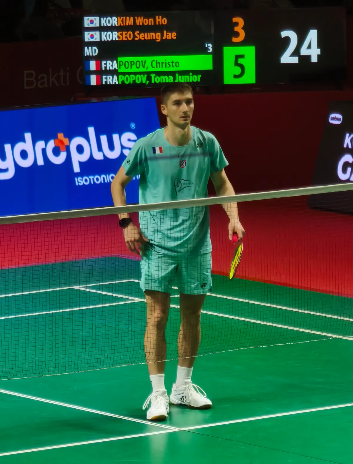 Un joueur de badminton en maillot vert d'eau, une raquette à la main, se tient debout sur un court vert derrière le filet, devant un tableau d'affichage de double messieurs.
